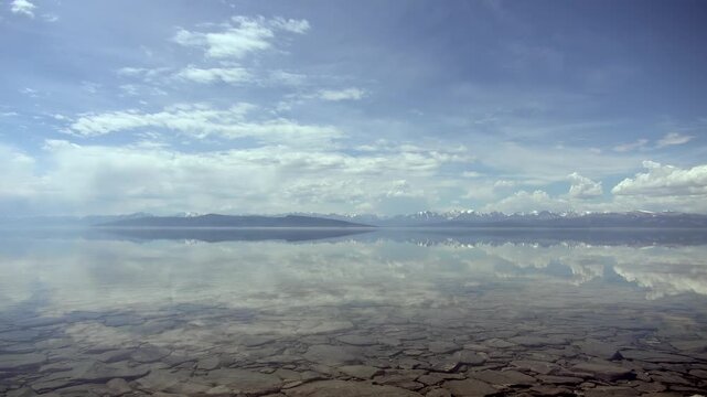 Clear Lake Surface Over Flat Rocks in Freshwater Shoreline Landscape. The view includes shallow transparent water exposes cracked stone patterns with pale clouds mirrored above.