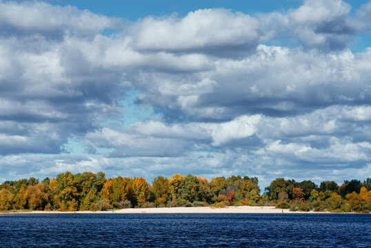 Autumn river scene with a sandy beach and vibrant fall foliage along the treeline, reflected in deep blue water beneath layered dramatic clouds and patches of bright blue sky.