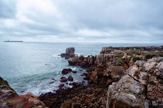Rocky coastal cliffs and rugged shoreline overlooking a calm ocean with a distant cargo ship on the horizon beneath a dramatic cloudy sky, evoking solitude, nature and coastal exploration.