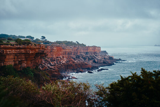 Dramatic sandstone cliffs drop to a rocky shoreline as waves lap the coast beneath an overcast sky, creating a moody, windswept seascape ideal for travel, nature, and landscape themes.