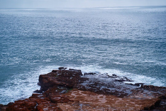 A rugged rocky coastline meets the blue ocean as waves break against the shore, creating foamy surf and tide pools; a serene, moody seascape conveying solitude, natural texture, and coastal erosion.