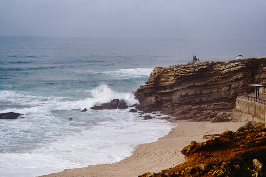 People stand on a rugged cliff watching waves crash onto a quiet sandy beach beneath an overcast sky, conveying coastal serenity, rugged nature and the power of the ocean coastline.