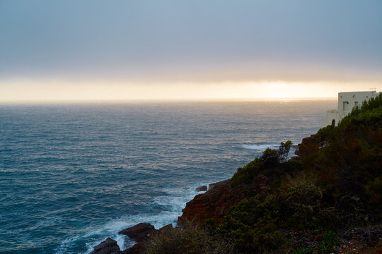 Solitary white house perched on a rocky cliff overlooking a vast ocean as golden light breaks the horizon. Moody sky, crashing waves and coastal vegetation convey peace, isolation and natural drama.