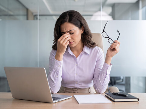 stressed white businesswoman sitting at modern office desk holding glasses and touching nose bridge for corporate workplace mental health banner ad