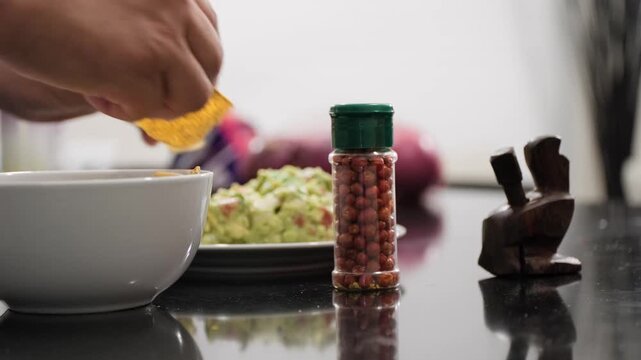A hand sets down a chiltepin pepper shaker before dipping tortilla chips into spicy homemade guacamole