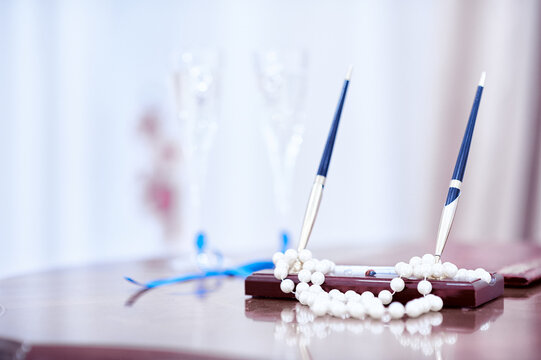 Elegant wedding signing set with two blue-and-silver pens and a strand of pearls resting on a polished table, champagne flutes blurred in the background evoking romance and celebration.