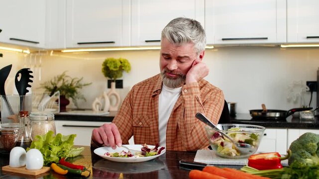 Middle-aged man grimacing while chewing vegetable salad in kitchen showing deep diet pressure and stress. Mature guy forcing healthy lunch for weight loss plan using strong willpower at home indoors.