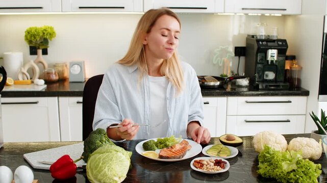 Middle-aged woman eating trout fish with avocado nuts and vegetables for keto diet in kitchen at home feeling satisfied. Young girl choosing low carb meal for healthy fit nutrition and weight loss.