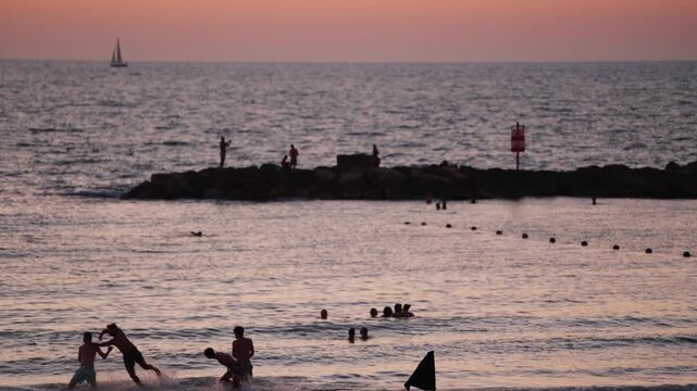 People bathing in the Mediterranean Sea at dusk in Tel Aviv, Israel. Summer holiday atmosphere as tourists and local enjoy fresh swim in the calm salt water. Sail boat and breakers in background