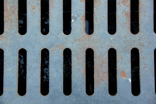 Full-frame top-down view of a weathered metal storm drain grate with vertical slots and rust spots over a street gutter.