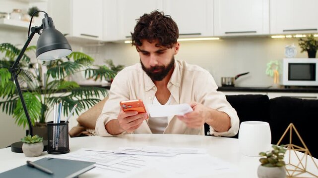 Indian man at home uses smartphone to enter bill amounts and calculate debt total feels worry. Arabian guy sitting at table surrounded by papers checks numbers again plans saving strategy for future.
