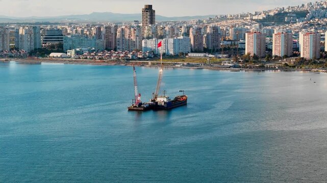 Aerial view of dredging operation near urban coastline with buildings behind. Crane barge and work vessel used for sediment removal in coastal water