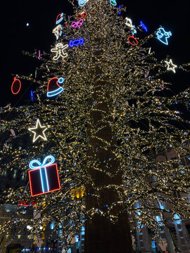 Large Christmas tree decorated with dense string lights and colorful neon ornaments stands outdoors at night