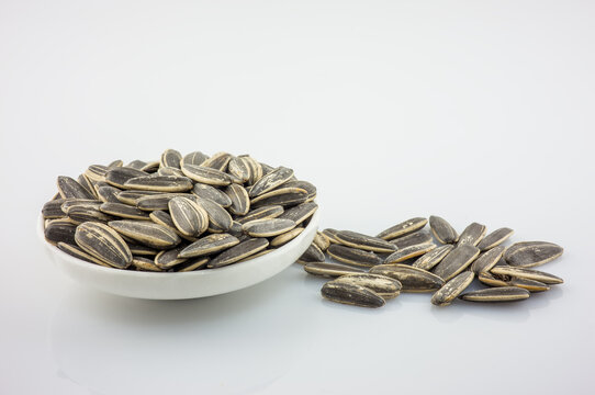 Striped sunflower seeds fill a white bowl with some scattered nearby, set against a plain light background.