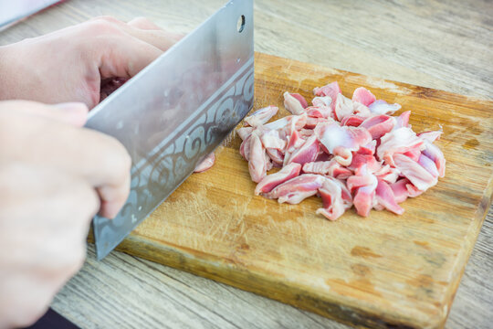 A person cuts fresh raw poultry gizzard into slices using a cleaver on a wooden cutting board for cooking preparation.