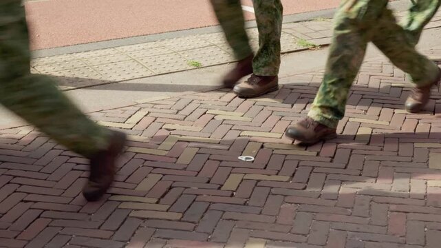 Low angle tracking view focuses on sturdy combat boots and camouflage pants of armed forces personnel marching over brick street during Oosterbeek memorial walk.