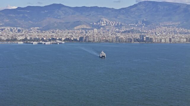 Passenger ferry crossing coastal bay with city skyline and mountains in background. Aerial view of public water transport in urban seascape