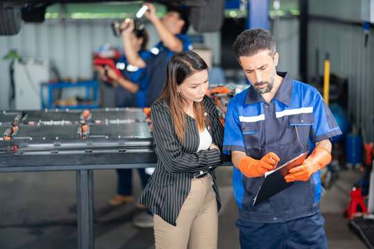 Customer at the garage listens as the service technician explains the battery report on his clipboard, her arms crossed.