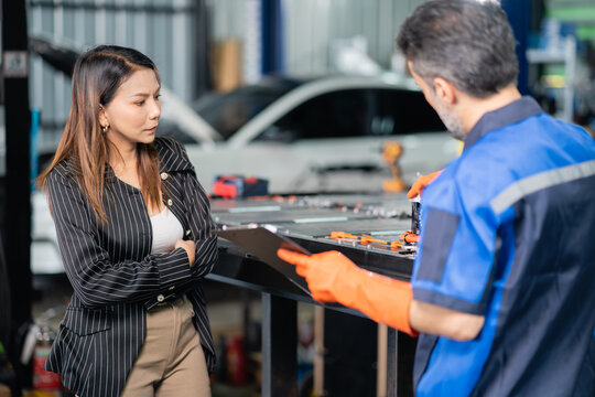 A customer at the garage discusses service with a technician holding a clipboard beside an open electric car battery pack.