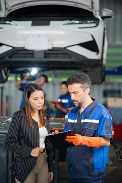 Customer and garage service technician share the clipboard under a raised electric car while colleagues inspect the underbody.