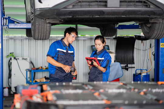 Technician holds a work light at waist level beside a colleague using a tablet beneath an electric car on a lift.