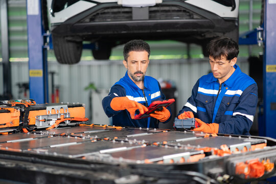 Beside an electric vehicle battery pack, a technician points at a tablet screen while a colleague holds an insulation tester.