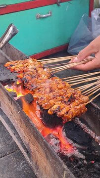 Hand grilling chicken satay skewers over charcoal fire. Indonesian street food, satay, barbecue, cooking, smoke, fire, UMKM, traditional, delicious.