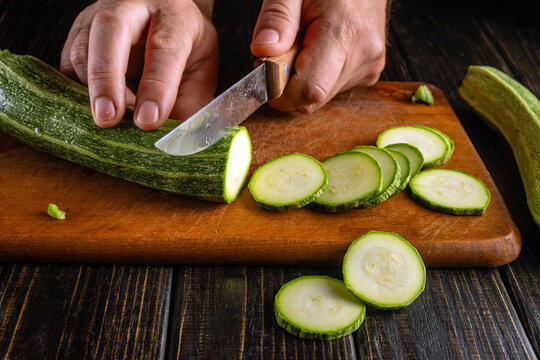 Hands prepare zucchini slices with sharp knife on cutting board. Vegetable is cut into even rounds, showing careful cooking prep and practical home kitchen scene with natural textures.