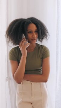 Woman talking on phone by window, standing with folded arms and listening during calm conversation. Everyday communication and personal assistance. Vertical video.