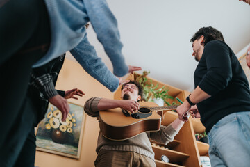 Fototapeta premium A young man plays an acoustic guitar while friends gather around him in a living room. The casual group sings and interacts during a relaxed home music session.