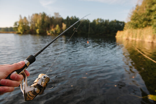 Close up view of an angler's hand holding a spinning fishing rod over a tranquil lake, enjoying outdoor recreation on Fisherman's Day from a boat, pier, or shore.