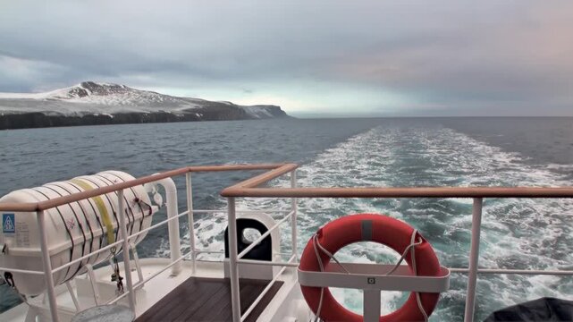 Ship Stern with Foamy Wake Moving Away from Snowy Coast in Cold Sea. The shot also shows how broad white wake runs from vessel stern across dark water with life ring across the visible setting.