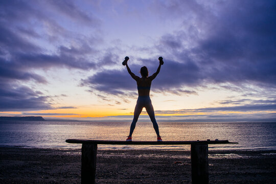 White woman exercising with dumbbells on beach at sunset in WA, USA