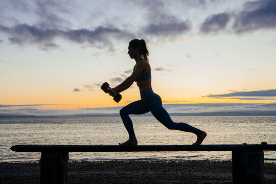 White woman exercising with dumbbells on bench at sunset by water