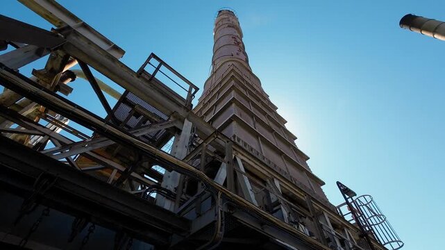 Industrial processing plant in backlight with massive chimneys, storage tanks, pipelines and complex machinery, showcasing large-scale production infrastructure under a bright blue sky.