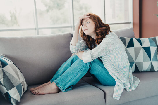 Pensive young woman sitting alone on sofa at home, looking worried and stressed in a quiet living room with natural light and casual clothing, feeling lonely and anxious