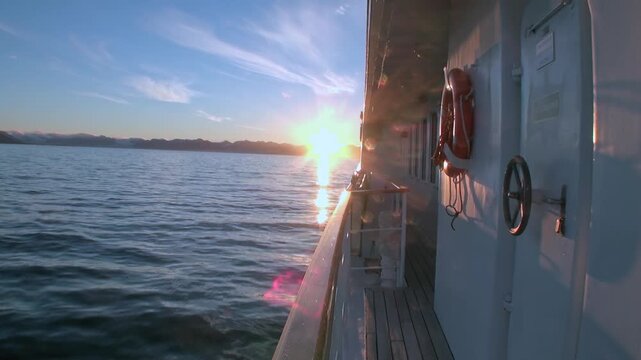 Vessel Side and Glowing Sea Moving beside Low Sun in Maritime Seascape. The shot contains gray ship wall and deck details border calm water with warm sun reflection and distant dark coast.