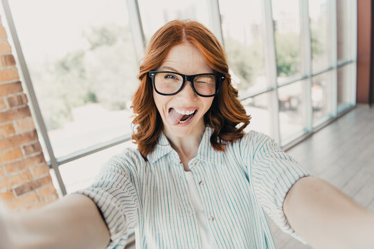 Playful redhead woman wearing glasses taking a funny selfie indoors, winking and sticking out her tongue in a bright modern office space