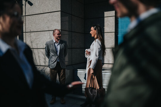 A man and woman in professional attire converse near a modern office building. Two blurred pedestrians pass in the foreground creating an urban workplace scene.