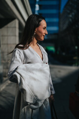 A stylish woman in a white coat stands on an urban sidewalk, looking to the side. The portrait captures a sunlit city moment with soft shadows and contemporary fashion. © qunica.com