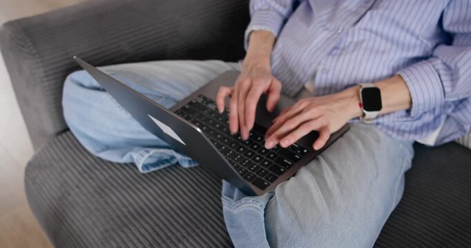 Close-up of female freelancer typing on laptop resting on her lap while sitting on sofa at home. Remote work in domestic setting