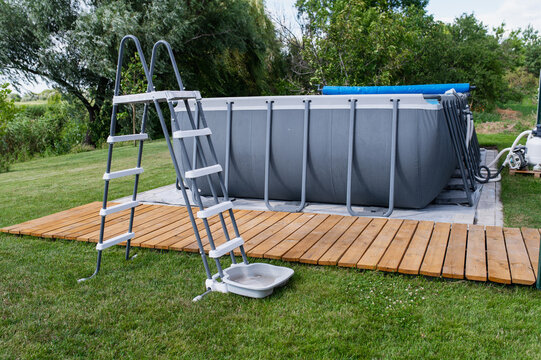 A frame pool with a ladder and a wooden deck on the lawn in the backyard relaxation area during summer; water treatment equipment, including a filter and pump.