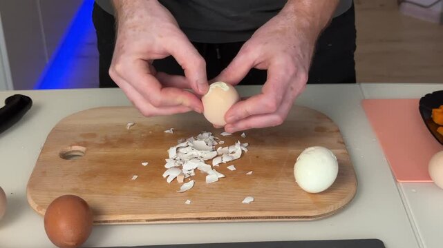 Overhead close-up of adult hands peeling hard-boiled egg, lifting brown eggshell to reveal smooth white egg and shell fragments on wooden cutting board kitchen prep