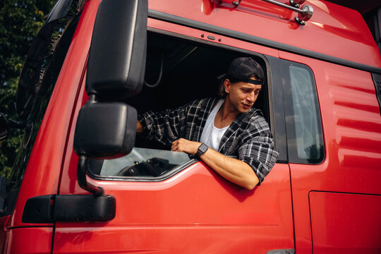 Young male truck driver wearing a plaid shirt leans out of the window of a red heavy-duty truck parked in a rural area with trees in the background