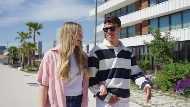 Young couple enjoying a sunny day stroll, linking arms on a modern walkway.