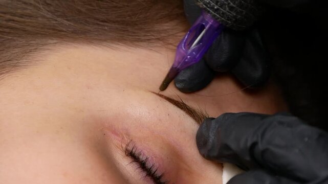 Eyebrow microblading procedure, specialist using precision pen to create defined brow shape. Detailed permanent makeup treatment focused on enhancing eyebrow appearance and symmetry