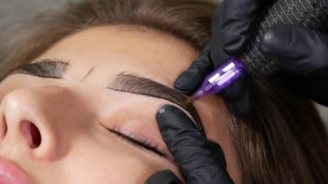 Specialist refining woman's eyebrow during microblading procedure, using precision tool to create defined brow shape. Permanent makeup treatment enhancing eyebrow symmetry and appearance