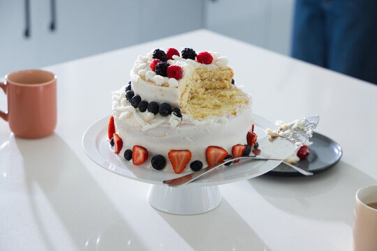 Two-tier white-frosted cake is sitting on pedestal stand on glossy counter, showing berry topping