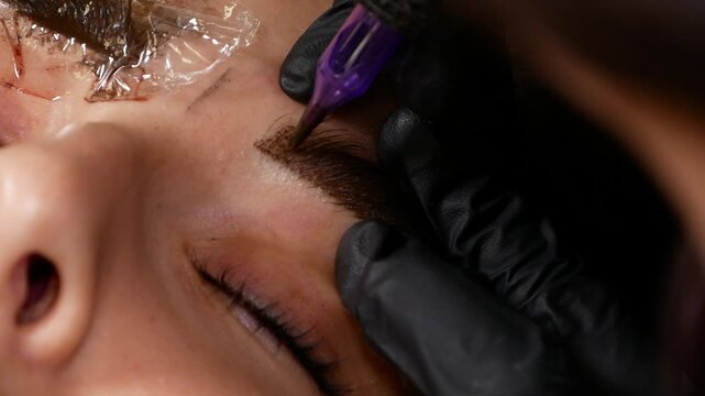 Specialist performing eyebrow microblading using precision pen, creating defined brow shape on woman's face. Permanent makeup procedure focused on eyebrow appearance and symmetry
