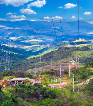 Power transmission towers and cables traversing a lush mountain landscape at a large mining operation in Minas Gerais, Brazil, under a bright blue sky.
